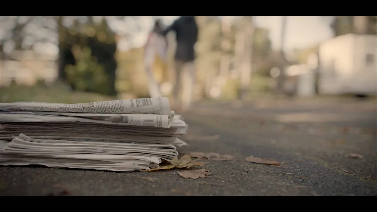 Close-up of a stacked bundle of folded newspapers on tarmac, with two blurred figures walking away on a suburban street beyond.