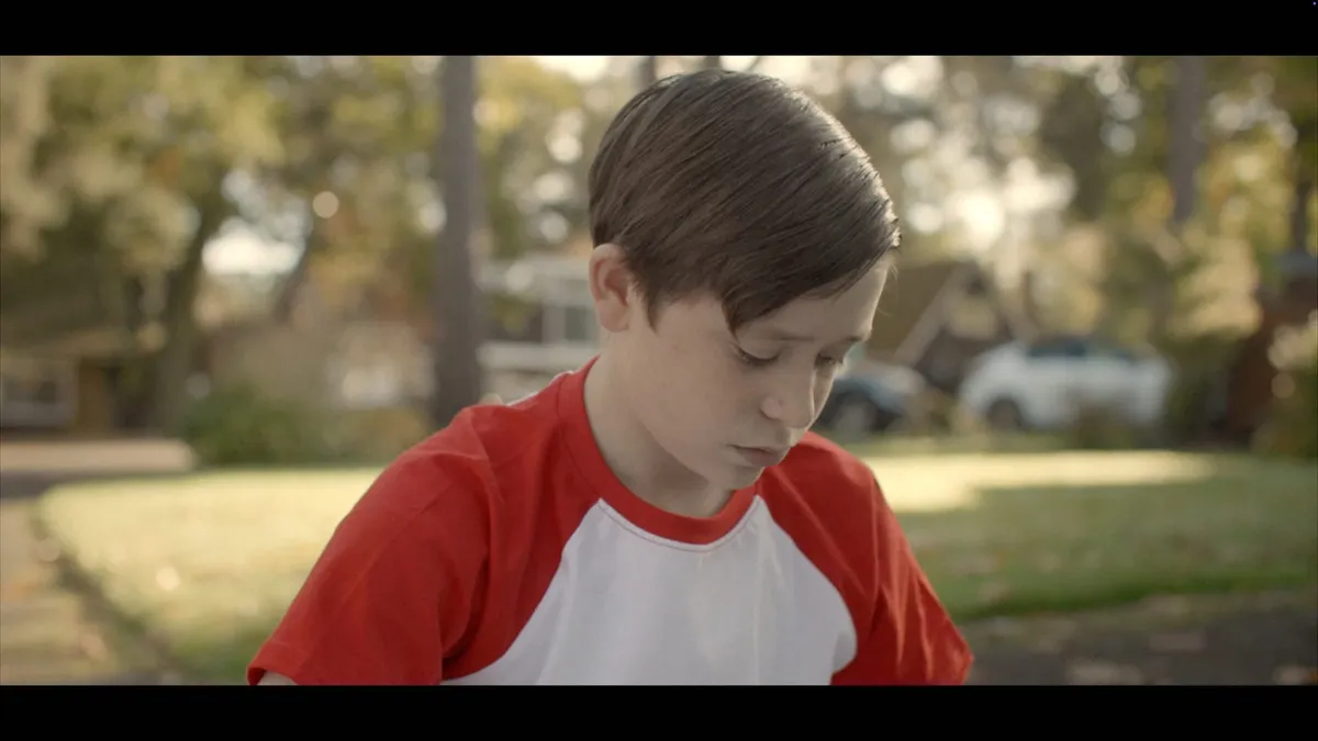 Close-up of a boy with neat brown hair, in a red and white raglan T-shirt, looking down on a suburban street with trees and cars blurred behind him.