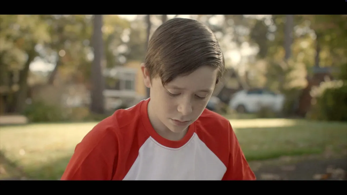 Close-up of a boy with neat brown hair, in a red and white raglan T-shirt, looking down, suburban street and trees blurred behind him.