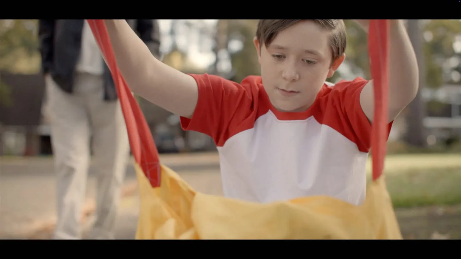Close-up of a boy with neat brown hair, in a red and white raglan T-shirt, holding open a yellow bag, an adult in beige trousers blurred behind him.