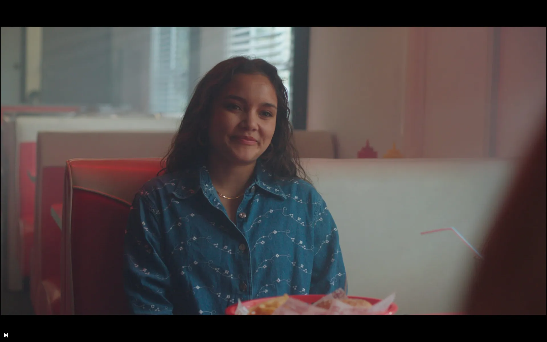 A woman sits in a diner booth in a denim shirt, tray of food in front, window light behind her.