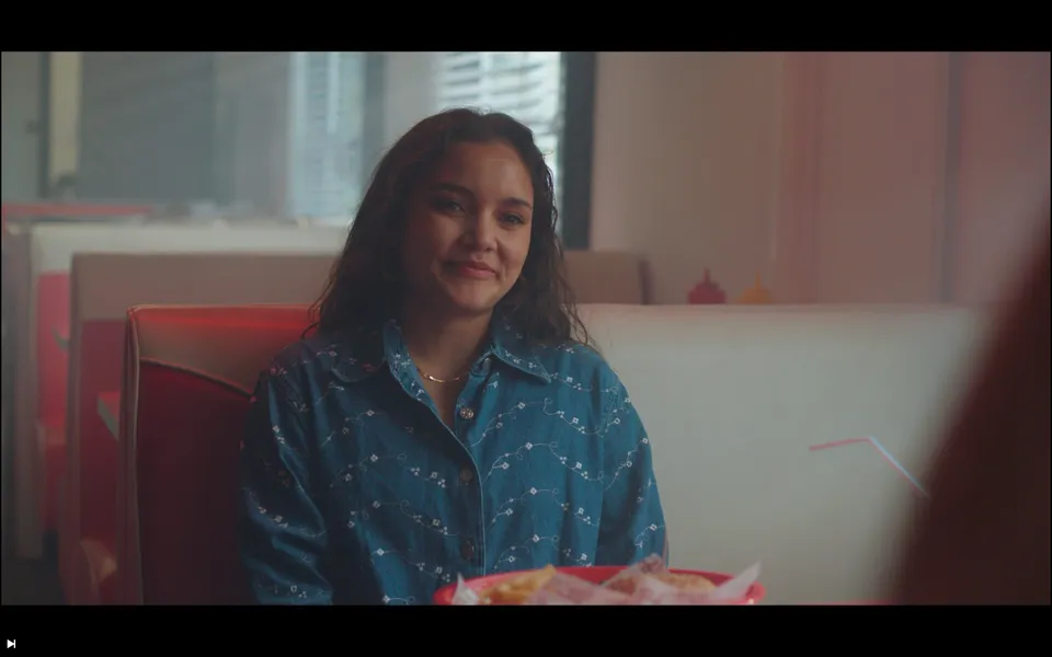 A woman sits in a diner booth in a denim shirt, tray of food in front, window light behind her.