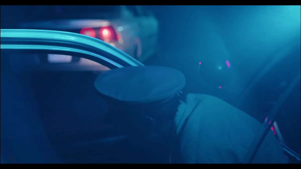 A police capped figure seen through a car window at night, another vehicle's rear lights glowing behind, lit in deep blue with red flare highlights.