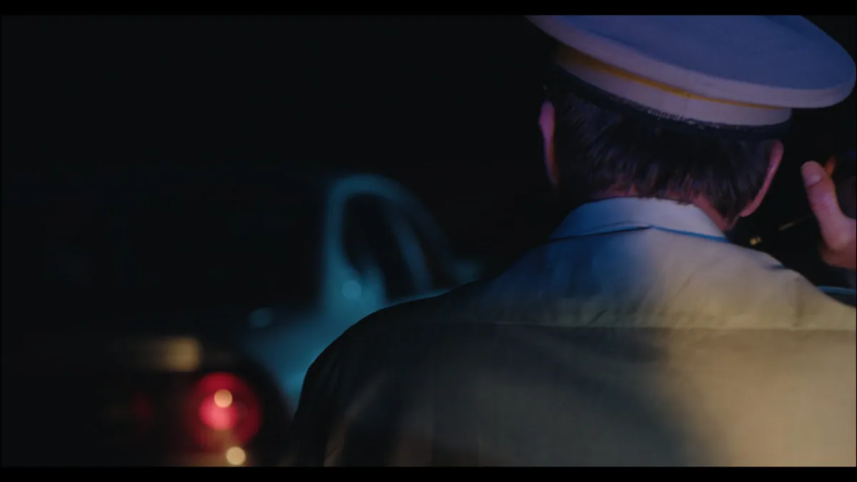Rear view of a man in a peaked police cap and shirt at night, a car's tail light glowing red in the background.