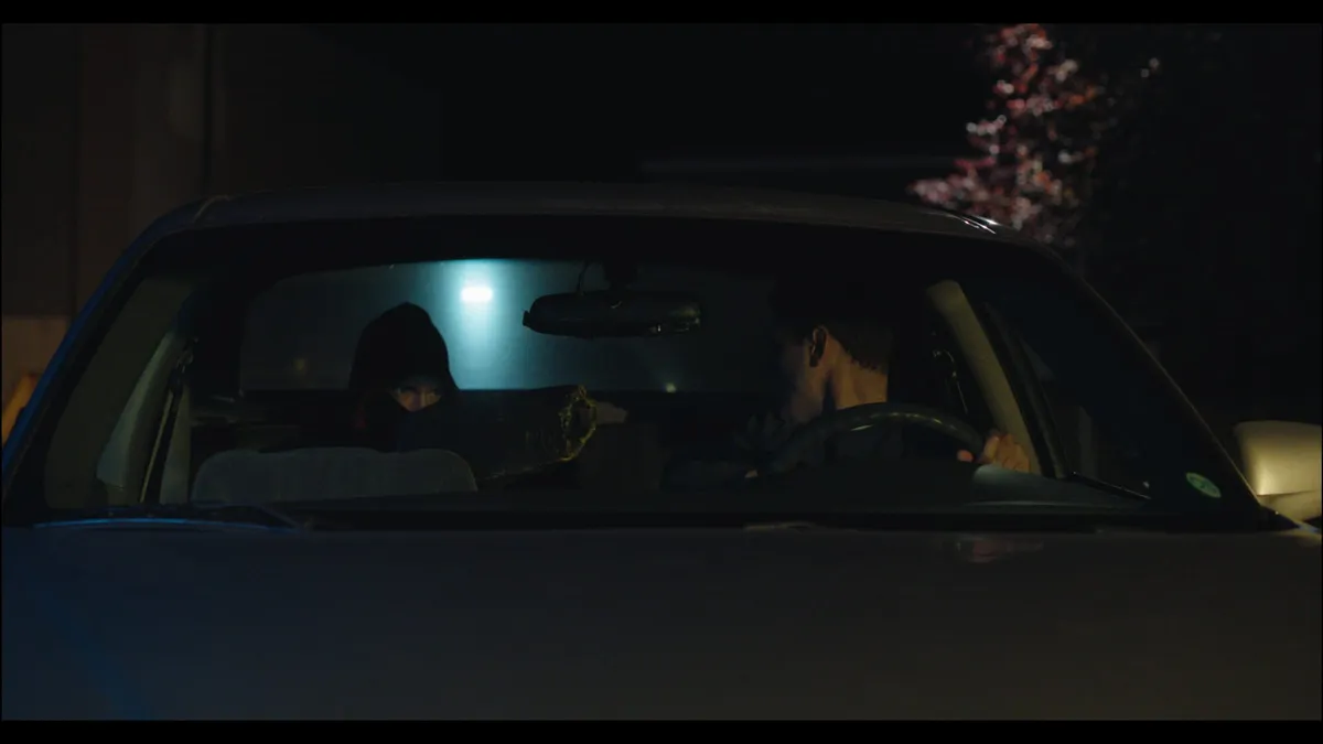 Night-time view through a car windscreen showing a driver and a hooded passenger in the back seat, lit by a dim interior light.