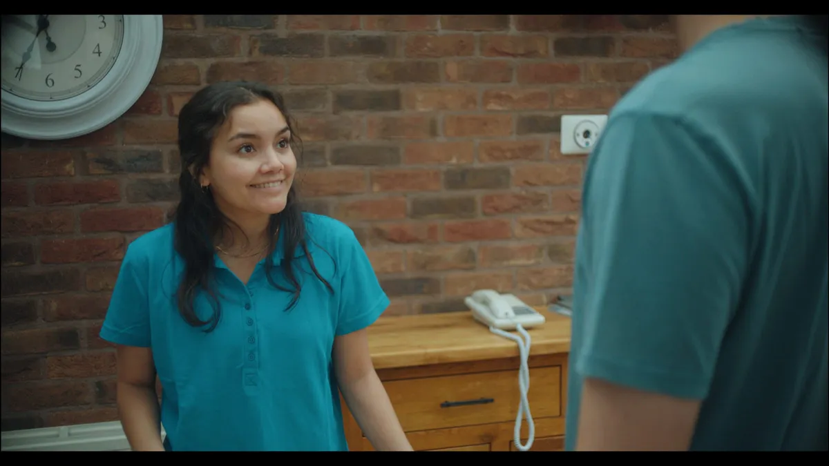 A woman in a teal polo shirt smiles at an unseen figure beside a brick wall, wall clock and wooden sideboard with telephone.