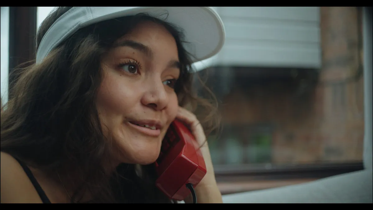 Close-up of a woman in a white visor holding a red corded telephone to her ear, shuttered window blurred behind her.