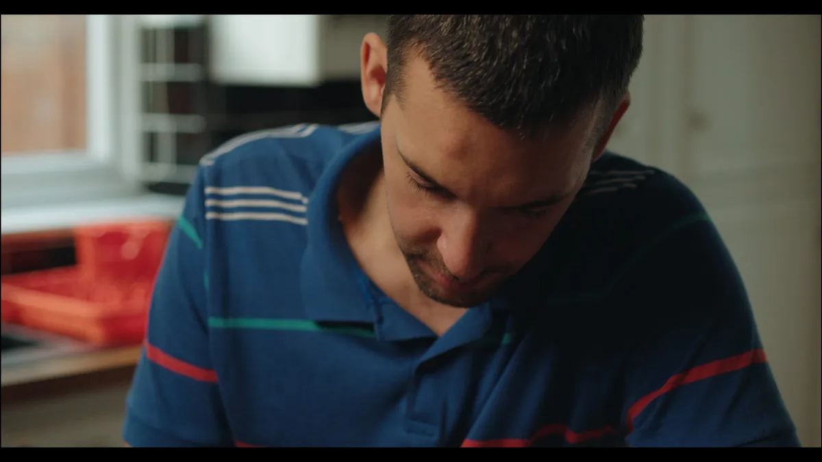 Close-up of a man in a striped polo shirt looking down, in a kitchen lit by daylight from a window behind him.