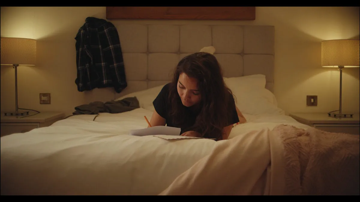A woman lies on a bed writing on paper, lit by two bedside lamps, a plaid shirt hanging on the headboard behind her.
