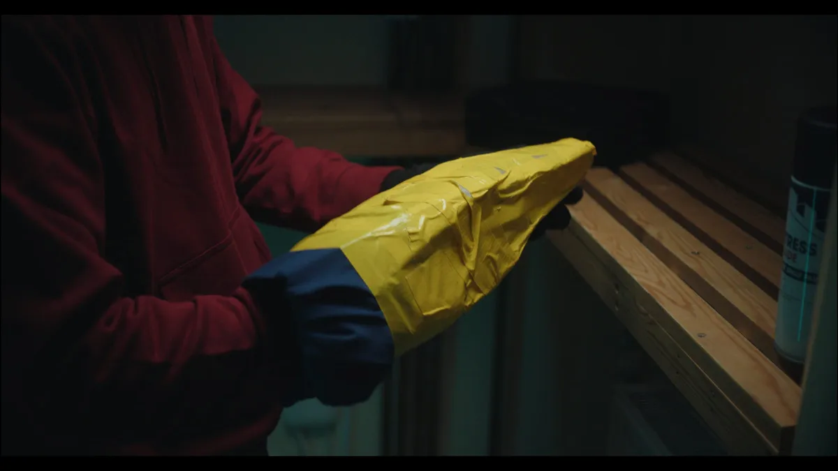 A gloved hand holds a yellow duct-taped object beside wooden shelving, dimly lit in a red-sleeved close-up.