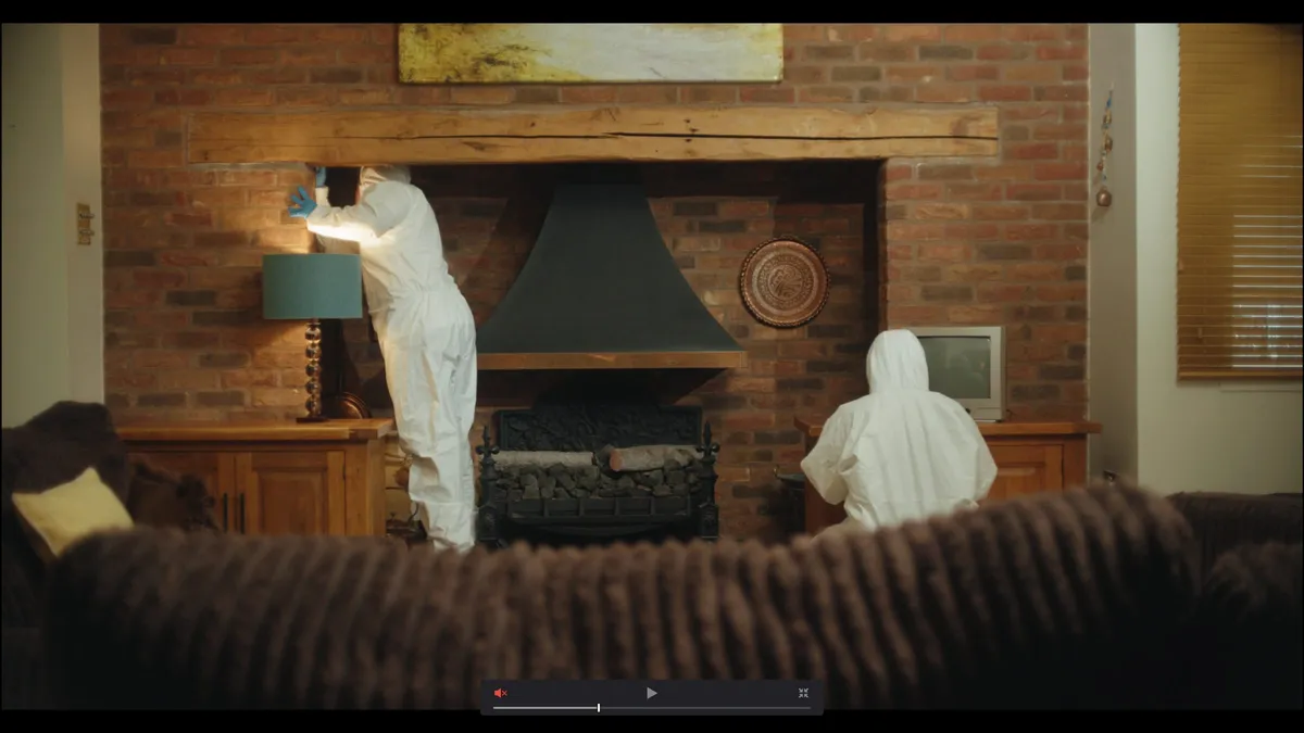 Two forensic investigators in white coveralls examine a brick fireplace and mantelpiece in a living room, viewed from behind a sofa.