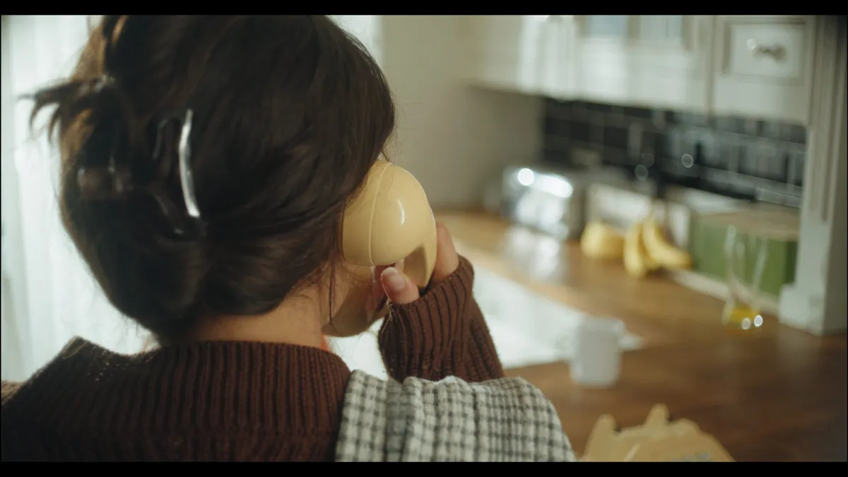 Rear view of a woman with hair in a clip holding a cream telephone receiver to her ear in a kitchen, bananas visible on worktop behind.