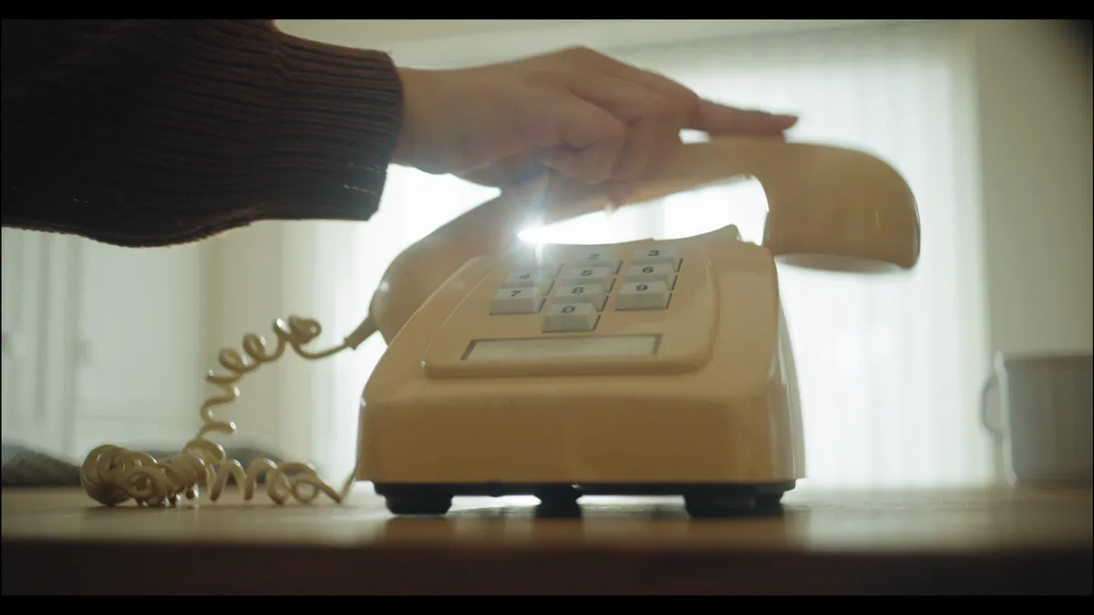 A hand lifts the receiver from a cream corded telephone on a table, backlit by a curtained window.