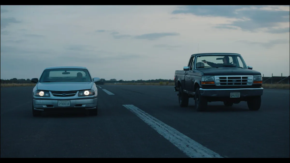 A saloon with headlights on faces a pickup on an open runway at dusk, a driver leaning out pointing a gun towards the car.