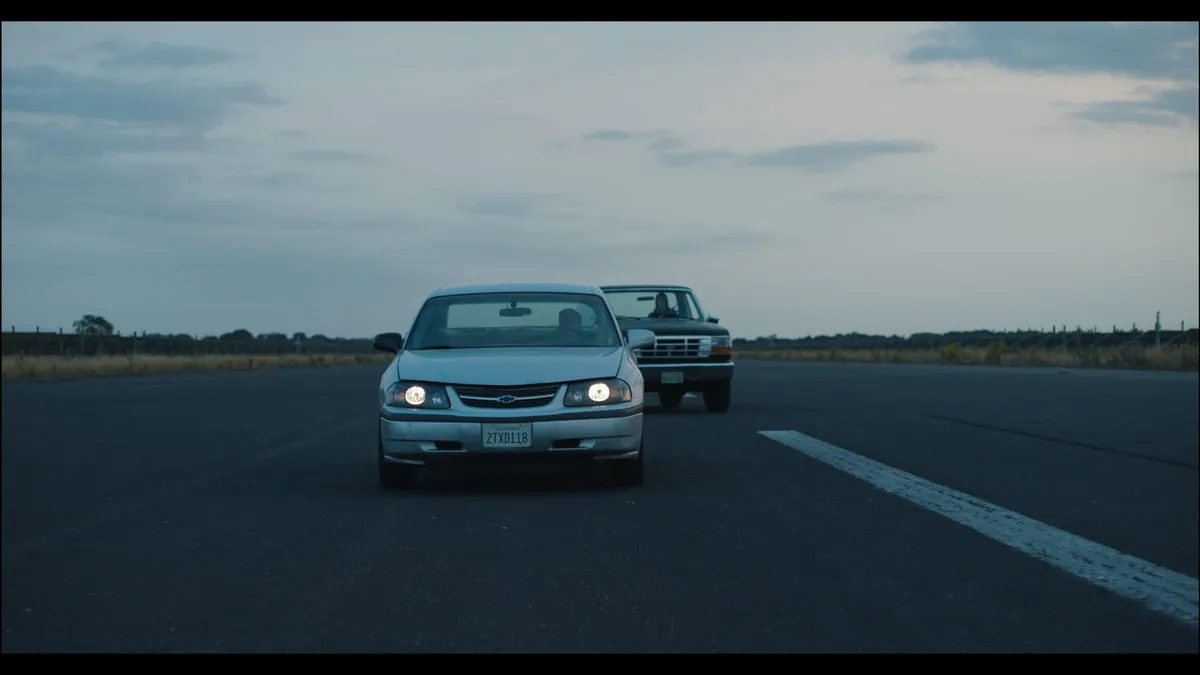 A saloon with headlights on and a pickup travel along an empty runway-like road under an overcast sky, framed from behind.