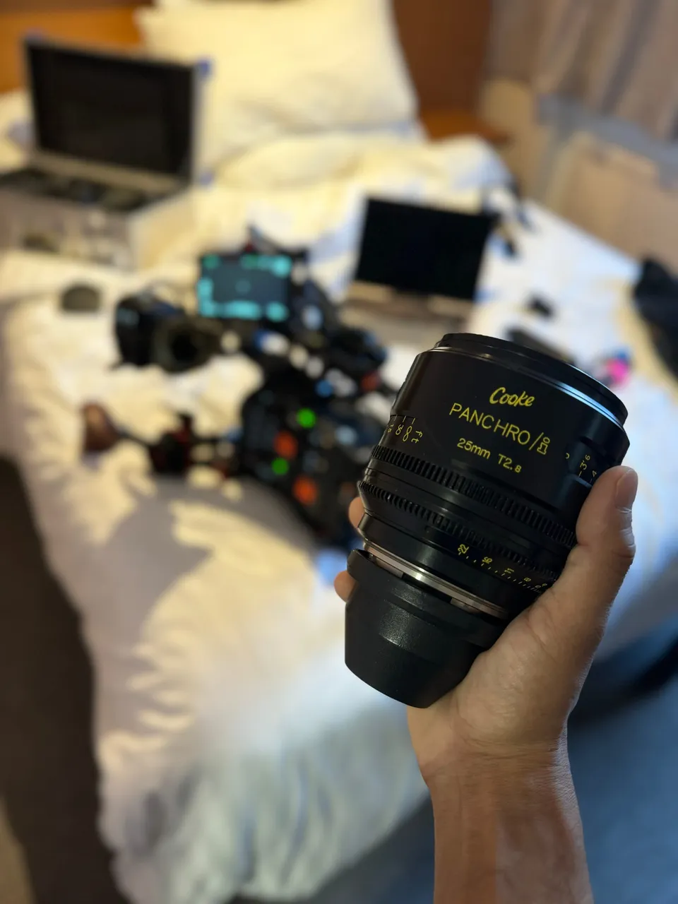 A hand holds a Cooke Panchro 25mm cine lens, with a camera rig and laptops visible out of focus on a bed behind.
