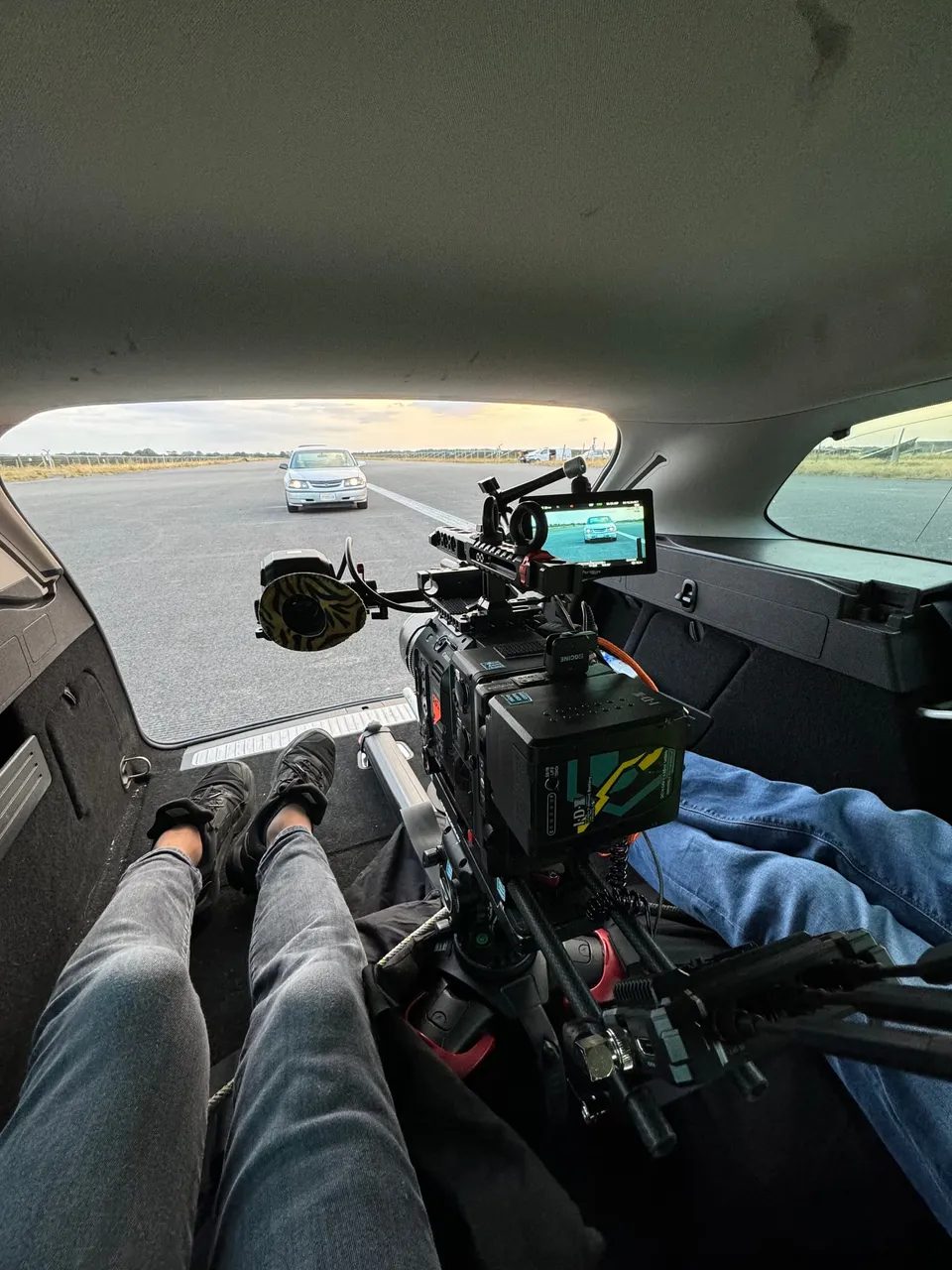 A camera crew member lies in a car boot operating a rigged cinema camera, framing a saloon car ahead on an open tarmac road.