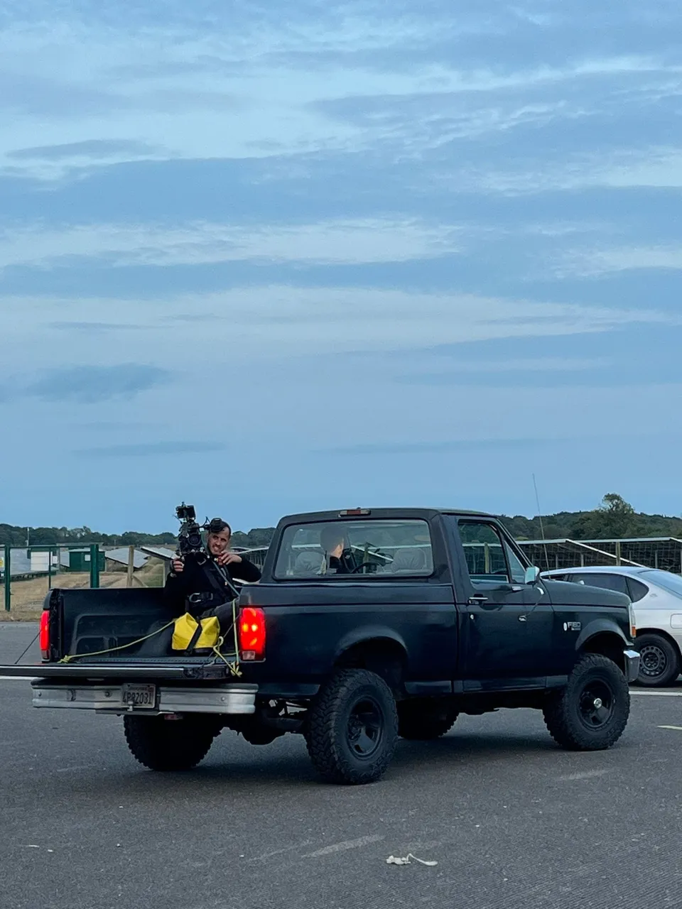 A camera operator crouches in the bed of a black pickup, filming with a shoulder-mounted camera, on a road under an overcast sky.
